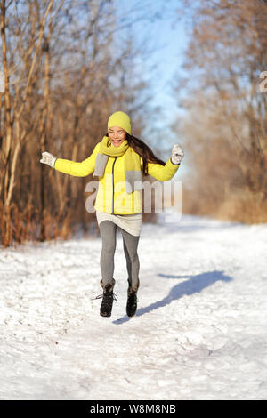 Glückliches Mädchen im Schnee Winter Landschaft läuft. Fröhliche junge asiatische Frau Spaß Wandern in der Natur forest park auf verschneiten Weg tragen gelbe Oberbekleidung mit warmen Zubehör: Stiefel, Handschuhe, Mütze, Schal. Stockfoto