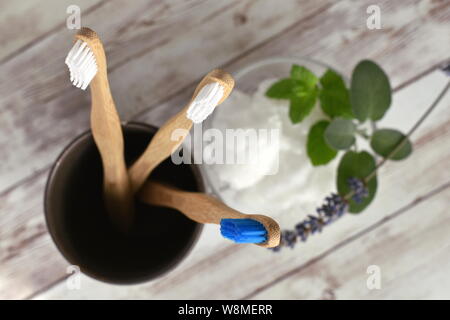 Blick von oben auf die drei Holz Bambus Zahnbürsten in Dunkelbraun Glas mit unscharfen Kokosfett im Glas dekoriert mit frischer Minze, Salbei und Lavendel Stockfoto