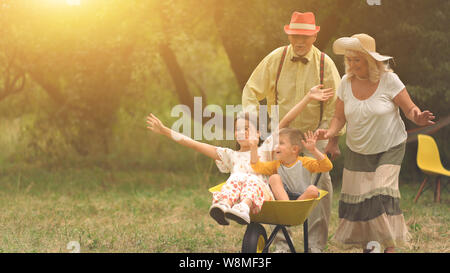 Oma und Opa drängen ihre Enkelkinder in einer Schubkarre 2 Stockfoto