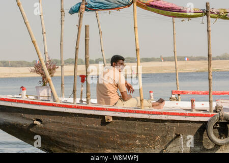 Einen indischen Mann sitzt auf einem hölzernen Boot auf dem Fluss Ganges in Varanasi, Uttar Pradesh, Indien, Südasien. Auch als Benares, Banaras und Kashi bekannt. Stockfoto