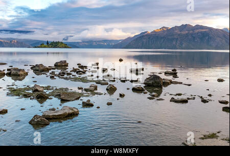 Ein Blick auf den Lake Wanaka am frühen Morgen, als die Sonne erhebt sich auf dem Hügel in der Ferne, vom Rand des Wassers, die Felsen in der foregrou Stockfoto