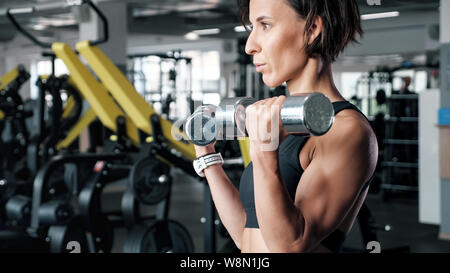 Portrait von athletischen reife Frau macht von reps eingestellte übung für Bizeps mit Hanteln in den Händen in der Turnhalle. Sie ist Hanteln. Ausbildung und Sport Konzept. Stockfoto