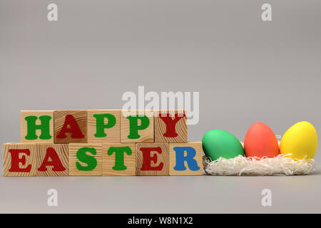 Drei Ostern bunte Eier im Nest liegen, neben der Holzwürfel mit dem Wort auf einem grauen Hintergrund glücklich. Frohe Ostern. Horizontale Fotografie Stockfoto