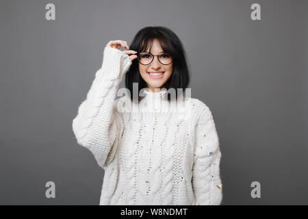 Hübsche junge brünette Frau in weißem Pullover Brillen tragen und lächelnd an der Kamera auf grauem Hintergrund Stockfoto