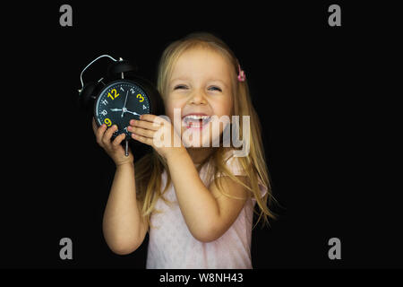 Zurück zum Konzept der Schule. Eine Schülerin mit einem Wecker in der Hand ist schockiert. Ein kleines Mädchen ist überrascht und sah mit einem schwarzen Uhr mit einer Hand h Stockfoto