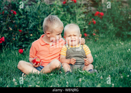 Zwei Brüder sitzen zusammen auf Gras im Park außerhalb gemeinsame Nutzung von Apple. Der ältere Bruder Umarmung beruhigen Sie sich beruhigen Schreien jüngeren wenig ein Kleinkind beruhigen. Sib Stockfoto