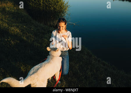 Glücklich lächelnde Frau, die auf dem Rasen See Bank und netten großen weißen Hund auf roten Leine in der Nähe Stockfoto