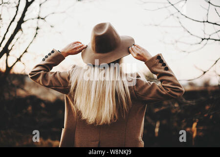 Rückansicht der jungen Dame in elegante Jacke berühren hat beim Stehen auf unscharfen Hintergrund der herbstlichen Landschaft. Elegante Frau berühren hat in der Natur Stockfoto