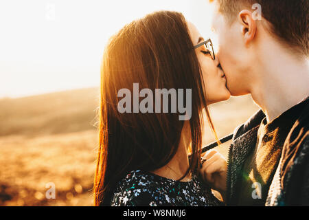 Attraktive junge Dame in modischen Brille küssen anonymer Freund in der Backe beim Stehen auf unscharfen Hintergrund der Landschaft auf sonnigen Tag. Frau ki Stockfoto