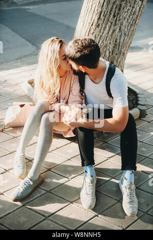 Romantische stilvolle Frau und bärtiger Mann sitzen auf Pflaster mit Baum hinter und Küssen im Sonnenlicht Stockfoto