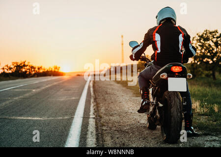 Rückansicht des Männlichen in Schutzhülle, Stiefel und Helm Reiten am Straßenrand bei Sonnenuntergang auf leere Autobahn hinterleuchteten Hintergrund Stockfoto