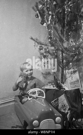 1950, historische, ein kleiner Junge seine Teddy-bären in seinem Spielzeug Fahrt sitzen umarmen - am Auto neben einem Weihnachtsbaum, England, UK. Stockfoto