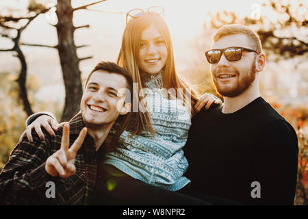 Schöne junge Frau lächelnd und Suchen an der Kamera, während zwischen zwei stattliche Kerle an einem sonnigen Tag in der Natur stehen. Hübsche Frau, die in der Nähe der Männer i Stockfoto