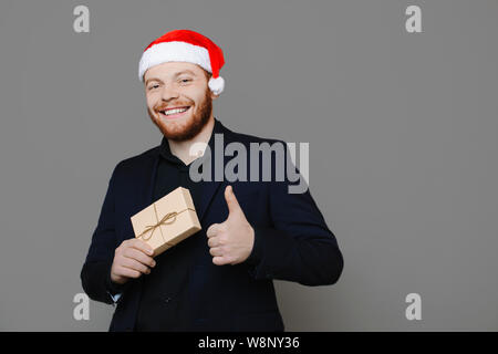Attraktiven jungen Mann im eleganten Anzug und Mütze holding Kleines präsent und zeigte mit dem Daumen-nach-oben-Geste beim Stehen auf grauen Hintergrund und cheerf Stockfoto