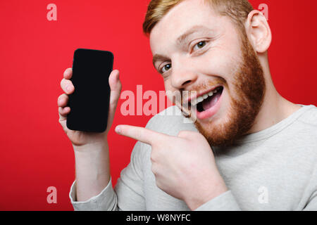 Stattlicher Mann mit Ingwer Bart zeigt Smartphone mit leeren Bildschirm und Kamera mit aufgeregten Gesichtsausdruck beim Stehen auf hellen re Stockfoto