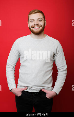 Attraktiven jungen Mann mit Ingwer Haar lächelnd und mit Blick auf die Kamera beim Stehen auf ein leuchtend roter Hintergrund und halten sich an den Händen in den Taschen der schwarzen Trou Stockfoto