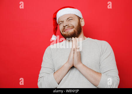 Hübscher Kerl in Weihnachten hat die gefalteten Händen und geschlossenen Augen beim Stehen auf rotem Hintergrund und machen möchten Stockfoto