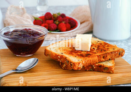 Zwei Toasts mit Stückchen Butter auf Holzbrett und Schüssel mit himbeermarmelade am Küchentisch. Stockfoto