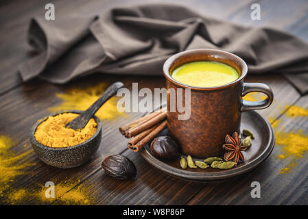 Traditionelle indische Gelbwurz Milch trinken ist golden Milch mit Zimt, Nelken, Pfeffer und Gelbwurz auf einer hölzernen Hintergrund mit Gewürzen Stockfoto