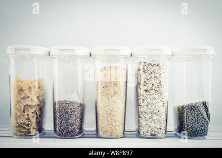 Verschiedene ungekochtem Getreide, Körner, Bohnen und Nudeln für gesundes Kochen in Kunststoffbehältern auf Holztisch, blauer Hintergrund, close-up. Die Lagerung von Lebensmitteln c Stockfoto