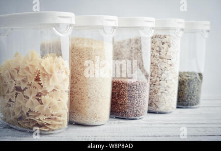 Verschiedene ungekochtem Getreide, Körner, Bohnen und Nudeln für gesundes Kochen in Kunststoffbehältern auf Holztisch, blauer Hintergrund, close-up. Die Lagerung von Lebensmitteln c Stockfoto