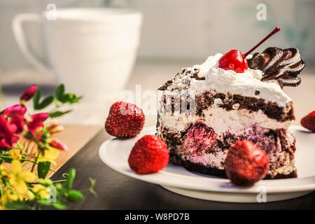 Ein Stück von einem Keks Kuchen mit Sahne und Kirschen liegt auf einem weißen Untertasse, mit Erdbeeren dekoriert. Neben der Tabelle ist ein Strauß von Wildblumen. Stockfoto