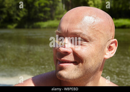 Unbehaarte kaukasischen lächelnden Mann lag in der Nähe von Fluss und schöne Schmetterling setzte sich auf seine Nase. Stockfoto