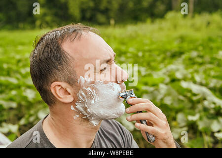 Weißer Mann mit Gesicht von Rasierschaum bedeckt ist mit Blade razor sein Gesicht auf die Natur im Wald zu rasieren. Stockfoto