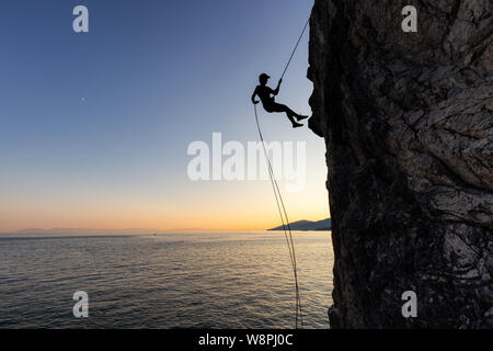 Silhouette einer Unkenntlich mann Abseilen nach einer steilen Klippe auf der rocly Ozean Küste während eines sonnigen Sommer Sonnenuntergang. In Lighthouse Park, West genommen Stockfoto