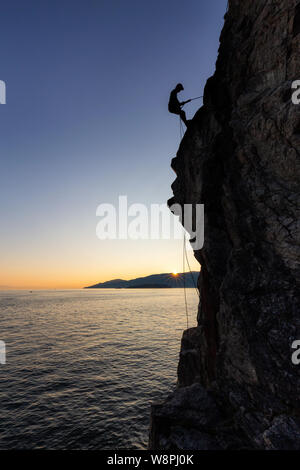 Silhouette einer Unkenntlich mann Abseilen nach einer steilen Klippe auf der rocly Ozean Küste während eines sonnigen Sommer Sonnenuntergang. In Lighthouse Park, West genommen Stockfoto