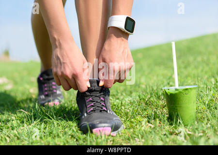 Laufschuhe, Sport Smartwatch und grünen Smoothie. Läuferin Schnürsenkel binden im Stadtpark, während das Trinken von einem gesunden Spinat und Gemüse Smoothie mit Smart watch Pulsmesser. Stockfoto