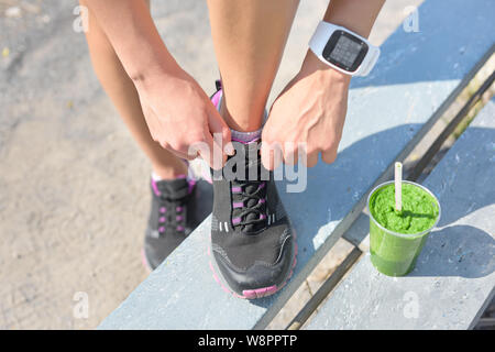 Laufschuhe, grünes Gemüse Smoothie und Sport erobert. Läuferin Schnürsenkel binden im Stadtpark, während das Trinken von einem gesunden Spinat und Gemüse Smoothie mit Smart watch Pulsmesser. Stockfoto