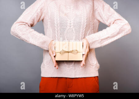 Die Ansicht der Frau in warmer Strickpullover und Terrakotta hose Holding in die Hände, die in kleinen Karton auf grauem Hintergrund Stockfoto