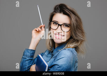 Seitenansicht attraktive junge freundliche Dame in Jean Shirt mit Notizblock und Stift an der Kamera schaut auf grauem Hintergrund Stockfoto