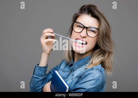 Seitenansicht attraktive junge Dame in Jean Shirt mit Notepad beißen, Stift und Kamera auf grauem Hintergrund Stockfoto