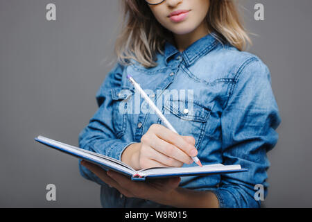 Die junge Dame in Jean shirt Notizen in Notepad mit Bleistift auf grauem Hintergrund Stockfoto