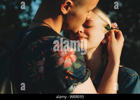 Erntegut Blick auf attraktiven Mann zärtlich an zierliche rosa Blume zu Haar hinter dem Ohr der reizende junge blonde Frau und umarmte ihn mit geschlossenen Augen Stockfoto