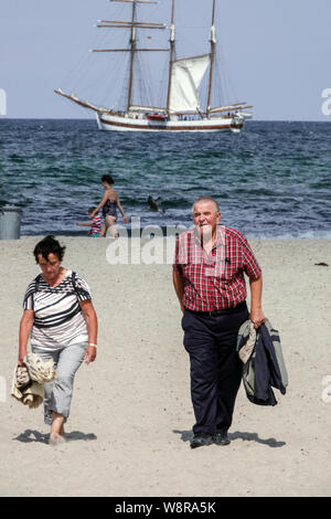 Seniorenpaar spazieren am Ostseestrand mit einem Hochschiff, das im Sommer in Warnemunde im Hintergrund segelt Stockfoto