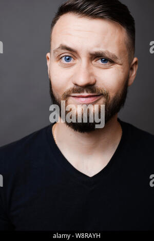 Gut aussehender bärtiger Kerl grinsend und die Augenbraue beim Stehen auf grauen Hintergrund und mit Blick auf die Kamera Stockfoto