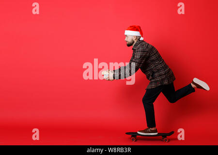 Seitenansicht des attraktiven Kerl in Mütze und casual Outfit tragen präsentiert und Reiten skateboard auf rotem Hintergrund Stockfoto