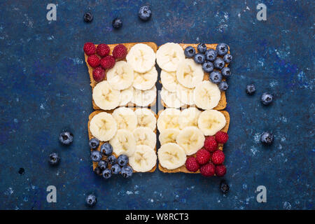 Gesunde Sandwich mit Erdnussbutter, Banane und Beeren auf einem dunkelblauen Tabelle. Ansicht von oben, flach Stockfoto