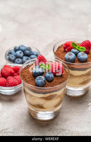 Classic Tiramisu Dessert mit Blaubeeren und Himbeeren in ein Glas und Schüsseln mit Beeren auf konkreten Hintergrund Stockfoto
