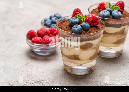 Classic Tiramisu Dessert mit Blaubeeren und Himbeeren in ein Glas und Schüsseln mit Beeren auf konkreten Hintergrund Stockfoto