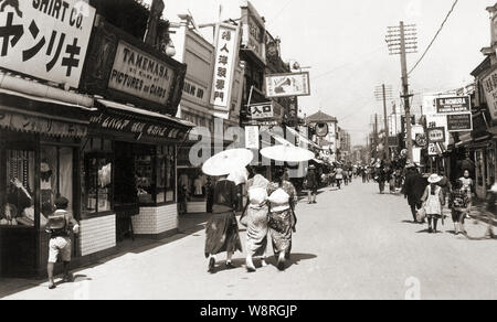 [1930er Jahre Japan - Japanische Einkaufsstraße in Kobe] - Käufer zu Fuß vor den Geschäften an Motomachi Itchome in Kobe, Hyogo Präfektur. Auf der linken Seite ist das Kobe Zweig der Foto Händler- und Portraitfotograf Torazo Tamemasa (1871-?). Tamemasa lief ein erfolgreiches Geschäft in Nagasaki, seit den 1890er Jahren. Die kobe Zweig wurde um 1899 (Meiji 32) eröffnet. Das 3-stöckige Gebäude im Hintergrund ist das berühmte Western style shirt shop Yamatoya. 20. Jahrhundert vintage Silbergelatineabzug. Stockfoto