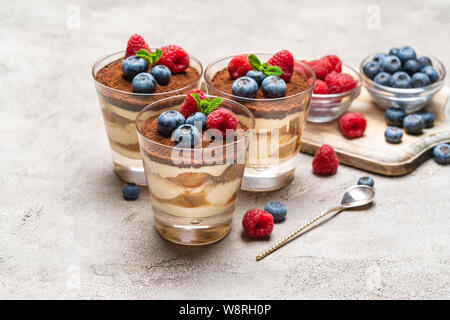 Classic Tiramisu Dessert mit Blaubeeren und Himbeeren in ein Glas und Schüsseln mit Beeren auf konkreten Hintergrund Stockfoto