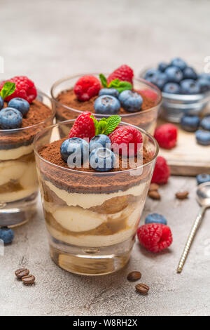 Classic Tiramisu Dessert mit Blaubeeren und Himbeeren in ein Glas und Schüsseln mit Beeren auf konkreten Hintergrund Stockfoto
