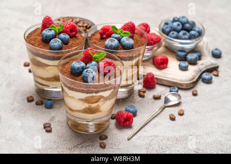 Classic Tiramisu Dessert mit Blaubeeren und Himbeeren in ein Glas und Schüsseln mit Beeren auf konkreten Hintergrund Stockfoto