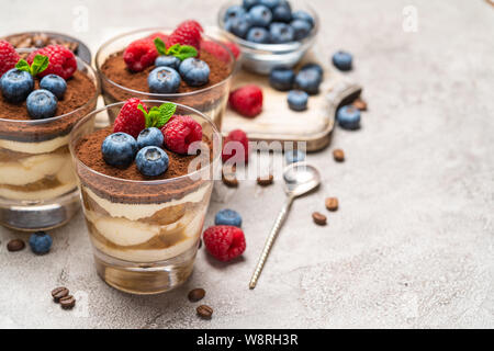 Classic Tiramisu Dessert mit Blaubeeren und Himbeeren in ein Glas und Schüsseln mit Beeren auf konkreten Hintergrund Stockfoto