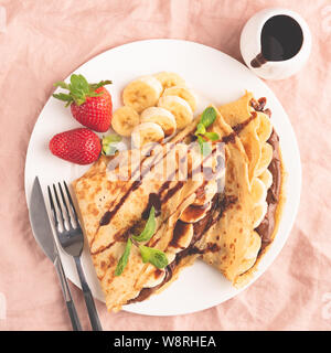 Französische Crêpe mit Schokolade und Banane auf dem Teller, rosa Hintergrund. Ansicht von oben. Leckere süße Speisen, Nachtisch Stockfoto