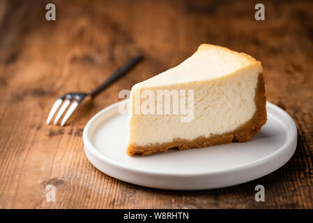 Käsekuchen Slice, New York Style klassische Käse Kuchen auf Holz- Hintergrund. Scheibe der leckere Kuchen auf weissem Teller mit Dessert Gabel Stockfoto